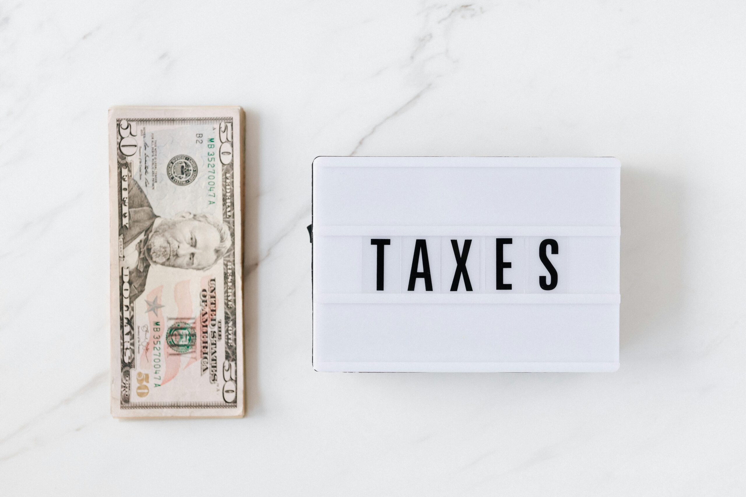 pexels-photo-4386369-4386369 Overhead view of a fifty-dollar bill and a lightbox with 'TAXES' on a marble surface.